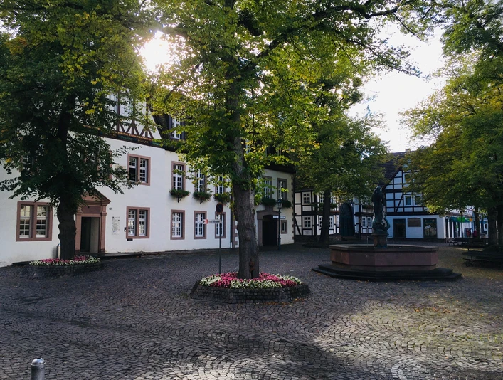 Kellerplatz in Beverungen Der Kellerplatz in Beverungen zeigt ein weißes Rathaus mit Fachwerkgiebel. Ein Brunnen mit St.-Michaelsfigur steht im Mittelpunkt, umgeben von schattenspendenden Bäumen, die dem Platz eine einladende Atmosphäre verleihen. Diese Szene betont die historische und zugleich lebendige Präsenz des Ortes.