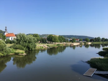 Das Foto zeigt den Verlauf des Weserflusses mit Uferbereich. Auf der linken Seite befindet sich eine weiße Kirche, umgeben von Bäumen, und im Hintergrund erstreckt sich ein bergiges Hinterland.