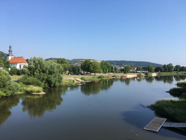 Das Foto zeigt den Verlauf des Weserflusses mit Uferbereich. Auf der linken Seite befindet sich eine weiße Kirche, umgeben von Bäumen, und im Hintergrund erstreckt sich ein bergiges Hinterland.