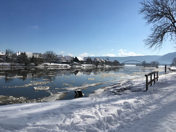 Das Foto zeigt die Weser im Winter bei Beverungen. Der Fluss ist teilweise mit Eisschollen bedeckt, im Schnee eingehüllt, und im Hintergrund ist die Weserbrücke zu sehen.