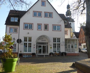 Großes, weißes Gebäude mit Arkadeneingang und Kopfsteinpflaster, im Hintergrund der Kirchturm der Katholischen Kirche.