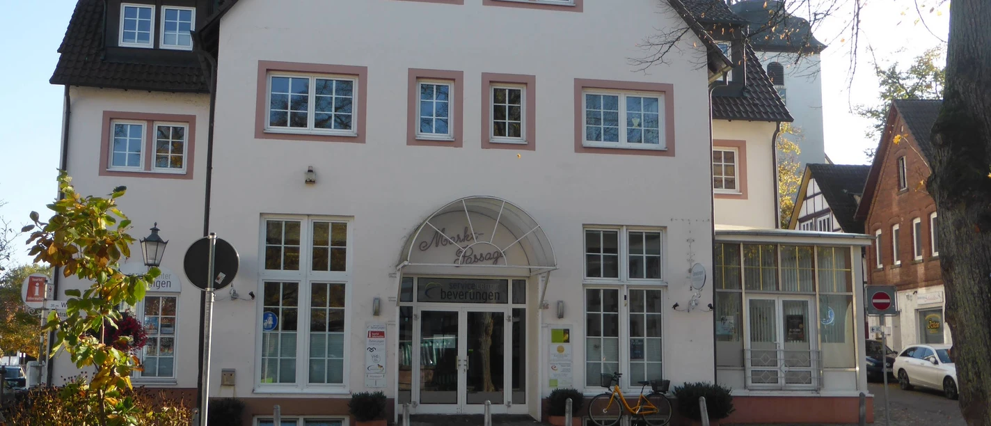 Großes, weißes Gebäude mit Arkadeneingang und Kopfsteinpflaster, im Hintergrund der Kirchturm der Katholischen Kirche.