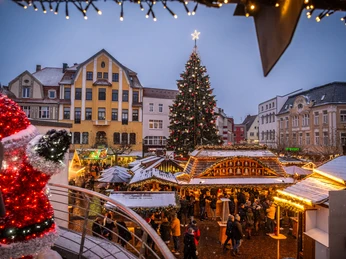 ProHerford_Weihnachtsmarkt2023_lowres_033.jpg Blick auf einen Weihnachtsmarkt mit geschmücktem Tannenbaum, Buden und flanierenden Menschen.