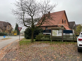 Parkplatz mit Laub, Bäumen und einem großen, rot-braunen Backsteingebäude, nahe einer Straßengabelung.