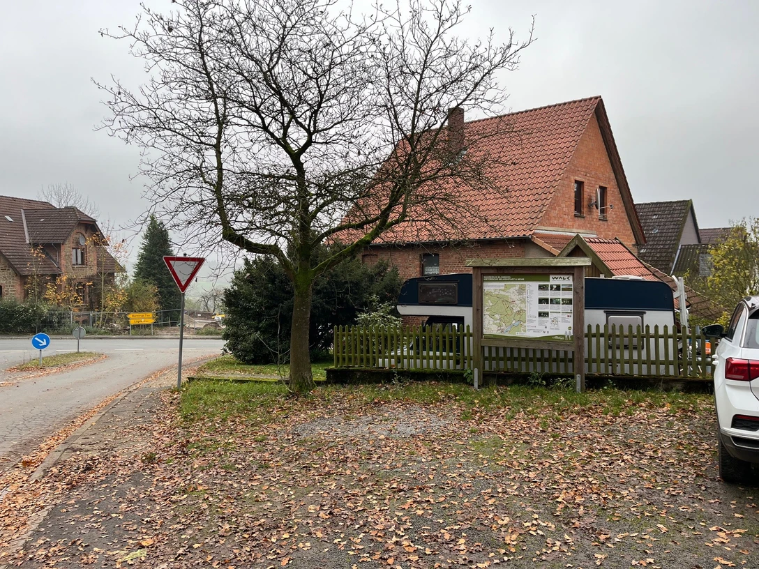 ParkplatzEschenbruch.jpg Parkplatz mit Laub, Bäumen und einem großen, rot-braunen Backsteingebäude, nahe einer Straßengabelung.