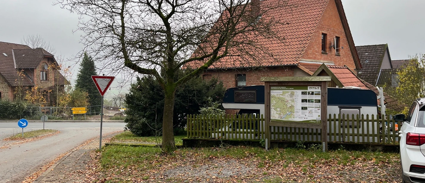 ParkplatzEschenbruch.jpg Parkplatz mit Laub, Bäumen und einem großen, rot-braunen Backsteingebäude, nahe einer Straßengabelung.