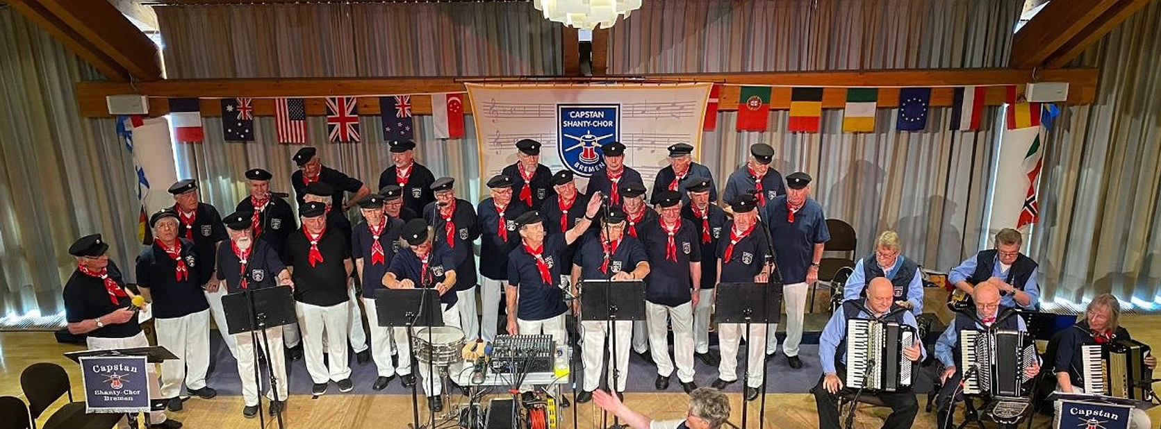 Capstan Shanty-Chor Der Capstan Shanty-Chor singt auf der Bühne, umgeben von Flaggen, mit Akkordeon-Begleitung.