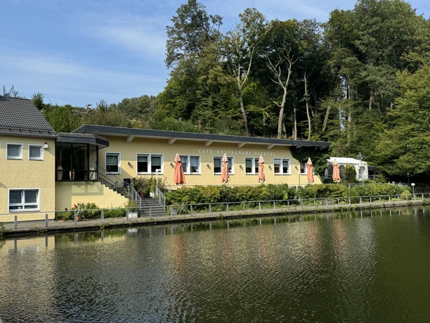 Café-Restaurant Uelfetal - Außenansicht Gelbes Gebäude am Seeufer mit Terrasse und orangenen Sonnenschirmen, umgeben von grünem Wald.