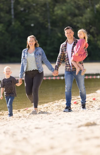 Badesee Hille Eine Familie spaziert an einem sandigen Ufer eines Sees entlang, umgeben von grünen Bäumen.