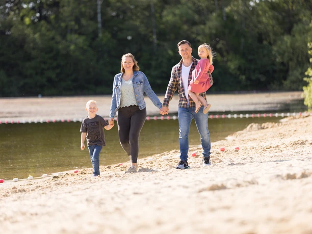 Badesee Hille Eine Familie spaziert an einem sandigen Ufer eines Sees entlang, umgeben von grünen Bäumen.