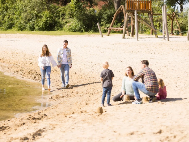 Familien und Paare genießen einen sonnigen Tag am Sandstrand eines ruhigen Sees mit Spielplatz.