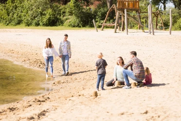 Badesee Hille Familien und Paare genießen einen sonnigen Tag am Sandstrand eines ruhigen Sees mit Spielplatz.