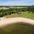 Luftaufnahme eines Badesees mit Sandstrand, umgeben von Bäumen und Wiesen, angrenzend ein Gebäude.