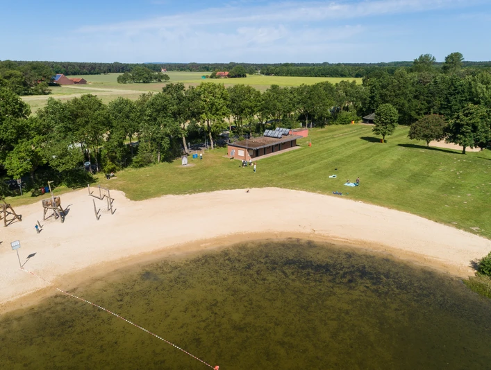 Luftaufnahme eines Badesees mit Sandstrand, umgeben von Bäumen und Wiesen, angrenzend ein Gebäude.