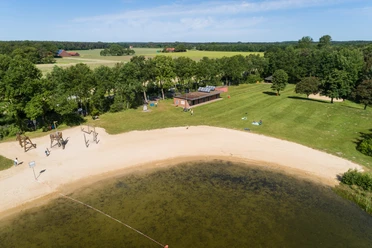 Badesee Hille Luftaufnahme eines Badesees mit Sandstrand, umgeben von Bäumen und Wiesen, angrenzend ein Gebäude.