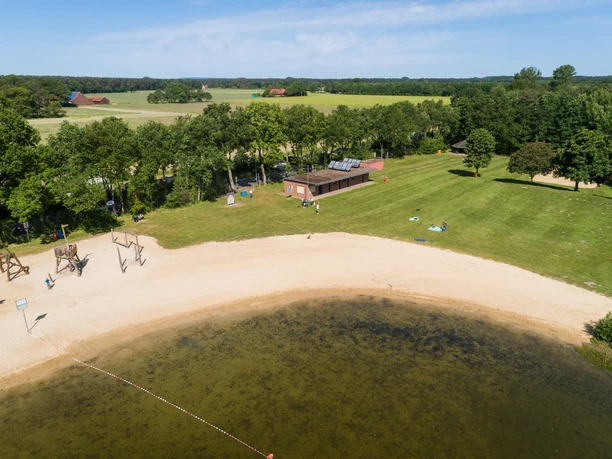 Badesee Hille Luftaufnahme eines Badesees mit Sandstrand, umgeben von Bäumen und Wiesen, angrenzend ein Gebäude.