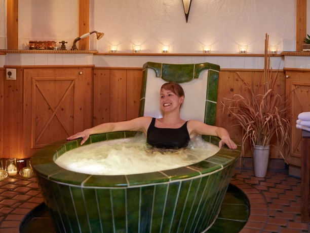 Sächsische Badestube Frau in schwarzem Badeanzug entspannt in einer grünen Keramikbadewanne, umgeben von Kerzenlicht und Holzwänden, in gemütlicher Atmosphäre.