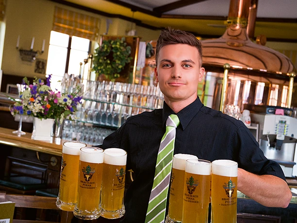 Číšník v černé košili a pruhované kravatě nese v útulném pivovaru s měděným kotlem v pozadí šest hrnků piva.