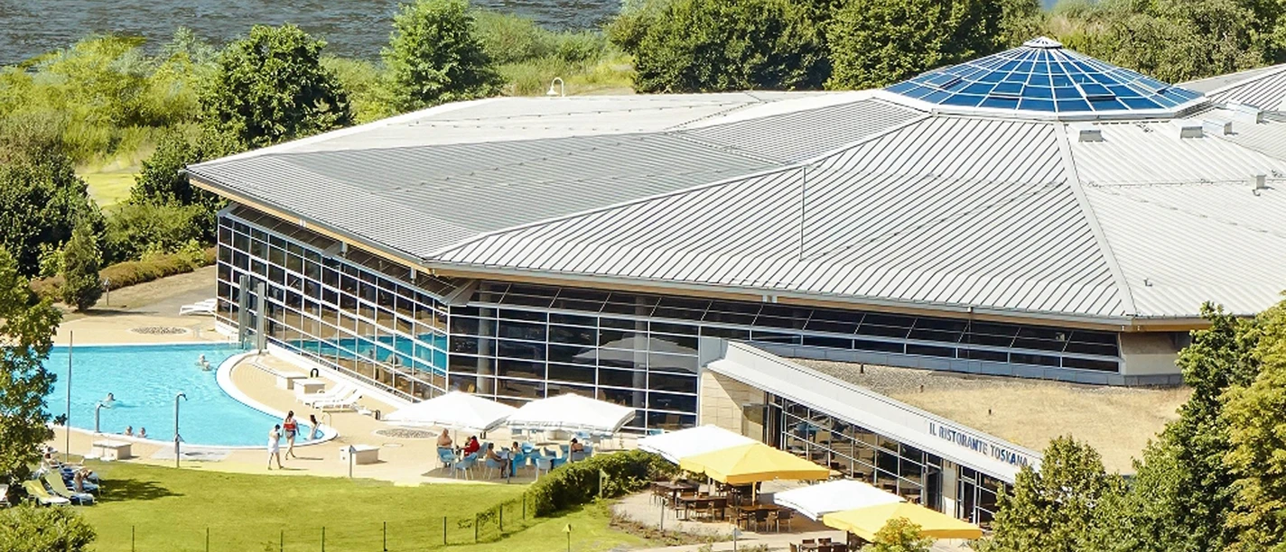 Modern gebouw met een groot glazen dak, omgeven door bomen, met een buitenzwembad en een terras met parasols; een rivier op de achtergrond.
