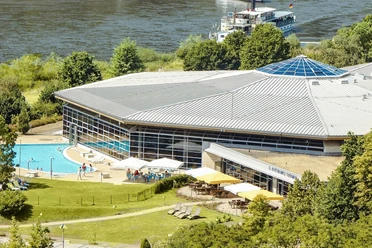 Il Ristorante Toskana Modernes Gebäude mit großem Glasdach, umgeben von Bäumen, mit einem Außenpool und einer Terrasse mit Sonnenschirmen; im Hintergrund ein Fluss.