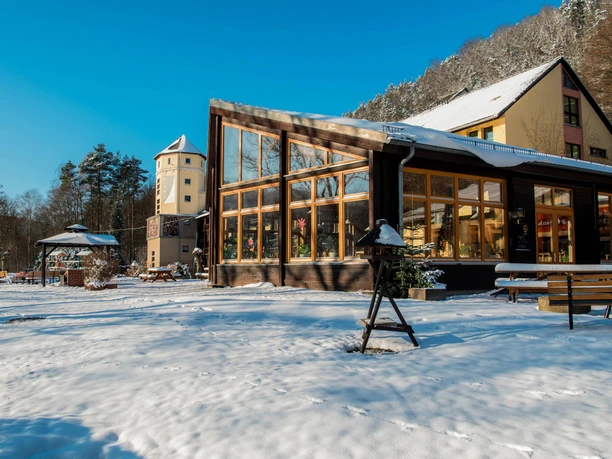 Een modern gebouw met grote ramen staat in een besneeuwd landschap, naast een toren en bomen onder een strakblauwe hemel.