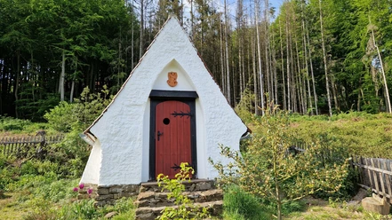 Lehmkapelle von Bodelschwingh Weiße Lehmkapelle mit roter Tür vor einem Wald, umgeben von einer Steinmauer und blühenden Pflanzen.