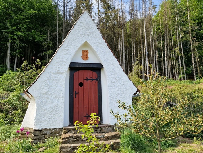 Lehmkapelle von Bodelschwingh Weiße Lehmkapelle mit roter Tür vor einem Wald, umgeben von einer Steinmauer und blühenden Pflanzen.