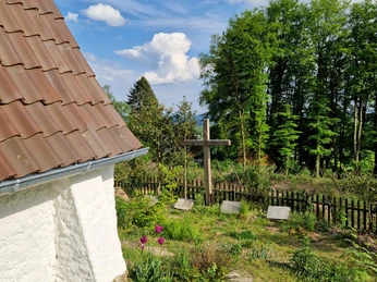 Lehmkapelle mit schrägem Dach, umgeben von Bäumen, Blumenbeeten und einem Holzkreuz im Garten.