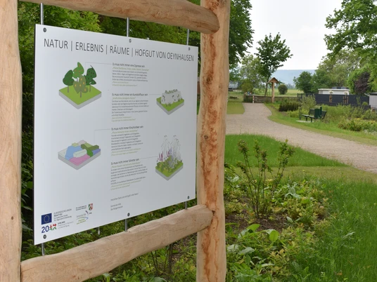 Informationsschild mit Text und Grafiken im Freien, umgeben von Naturwegen und üppigem Grün.