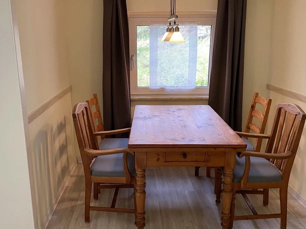 IMG_2912.jpeg Holztisch mit vier Stühlen in schlichtem Raum, Tageslicht durch Fenster mit blickdichtem Vorhang.