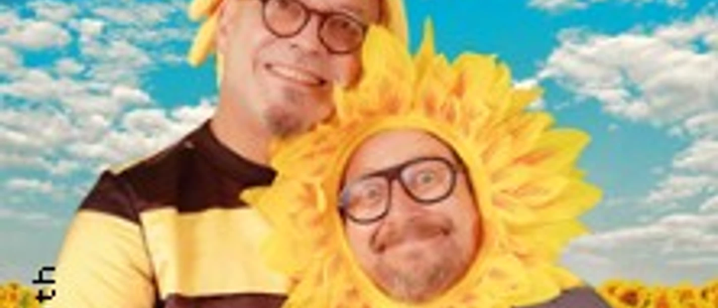 Zwei verkleidete Personen als Biene und Sonnenblume in einem Feld unter blauem Himmel.