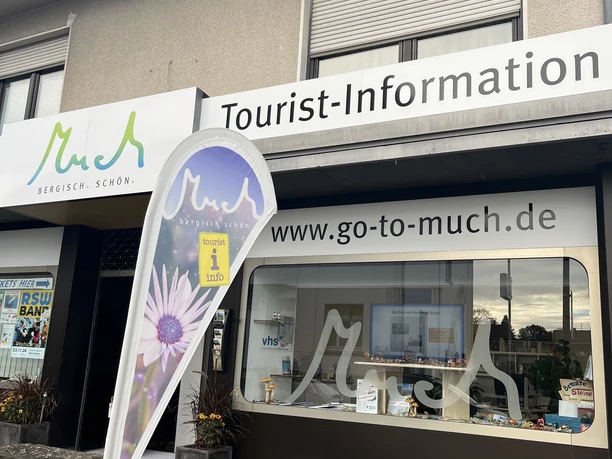 Tourist-Information Tourist-Information Much mit Fensterschild und Fahne, darunter Blumenbeet, für Besucheranfragen.