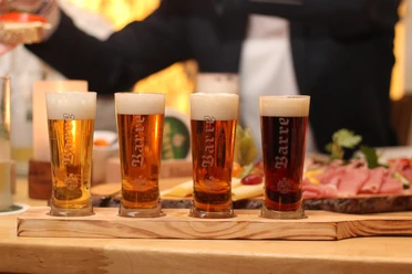Barres Brauwelt ProBier-Brett mit vier gefüllten Biergläsern, begleitet von rustikalem Imbiss auf Holz.