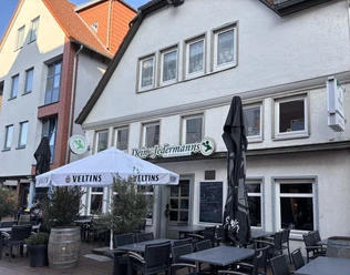 Jedermann_3.jpg Außenansicht eines Restaurants mit Terrasse, Holzmöbeln und Sonnenschirmen unter blauem Himmel.