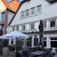 Jedermann_3.jpg Außenansicht eines Restaurants mit Terrasse, Holzmöbeln und Sonnenschirmen unter blauem Himmel.