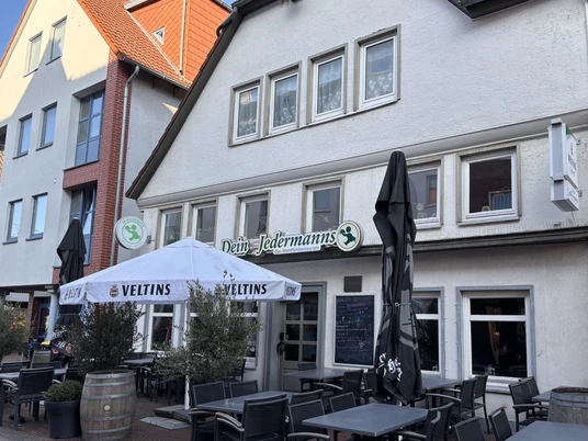 Jedermann_3.jpg Außenansicht eines Restaurants mit Terrasse, Holzmöbeln und Sonnenschirmen unter blauem Himmel.