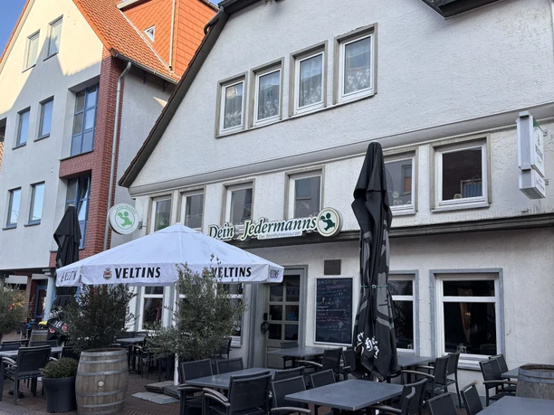 Jedermann_3.jpg Außenansicht eines Restaurants mit Terrasse, Holzmöbeln und Sonnenschirmen unter blauem Himmel.