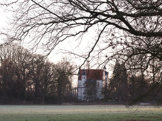 Ein großes, weißes Gebäude steht umgeben von kahlen Laubbäumen auf einer weitläufigen Wiese.