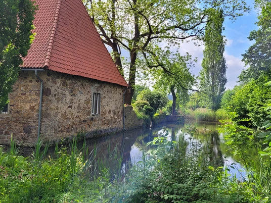 Gut Stockhausen Steinhaus mit rotem Dach am Teichufer, umgeben von üppigem Grün und Bäumen unter blauem Himmel.