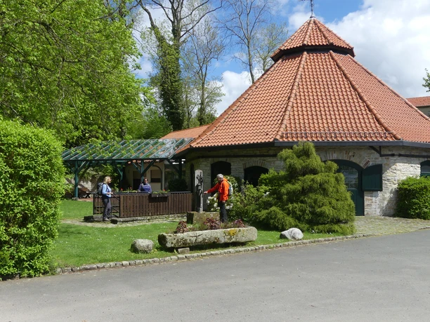 Uhdenhütte Runde Steinhütte mit rotem Ziegeldach in grüner Umgebung, Menschen unter Pavillon im Austausch.