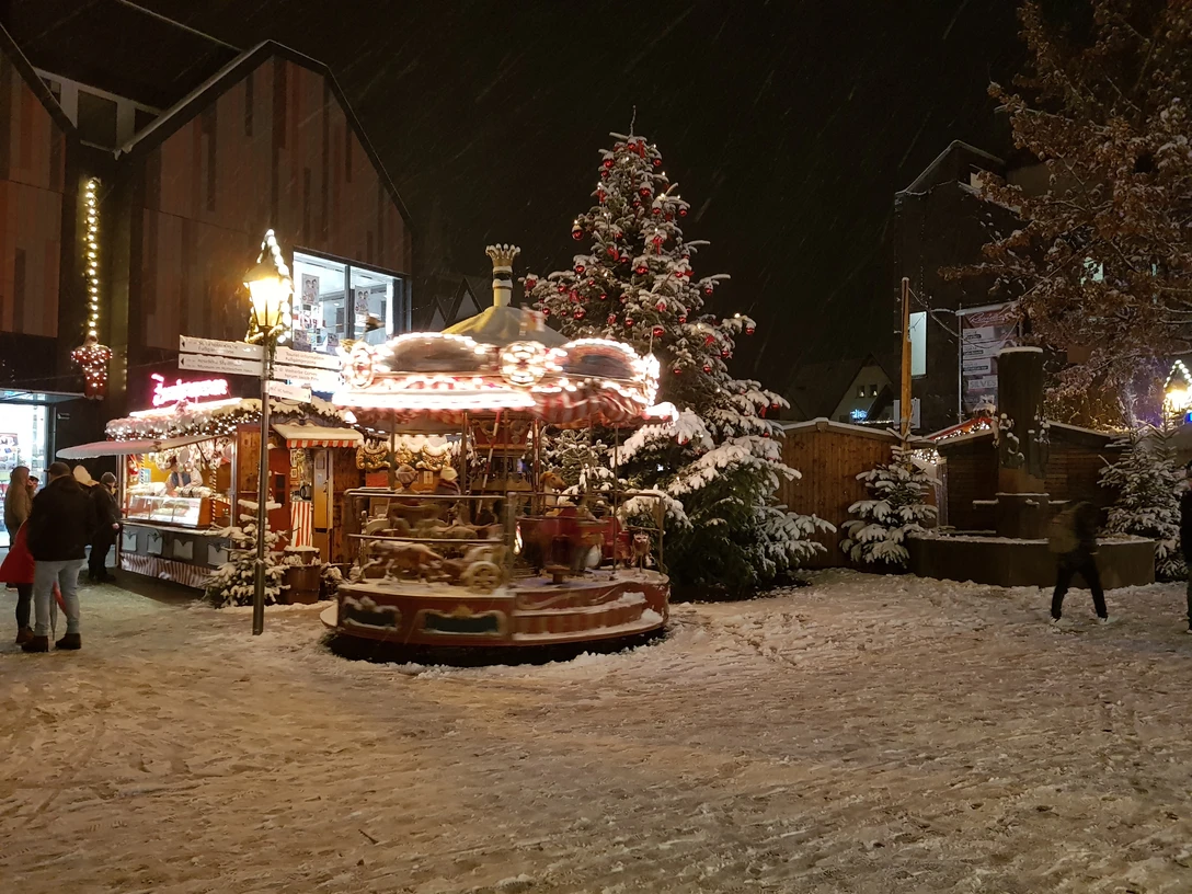 20171210_180126.jpg Ein verschneiter Weihnachtsmarkt am Abend mit beleuchtetem Karussell und geschmücktem Weihnachtsbaum.