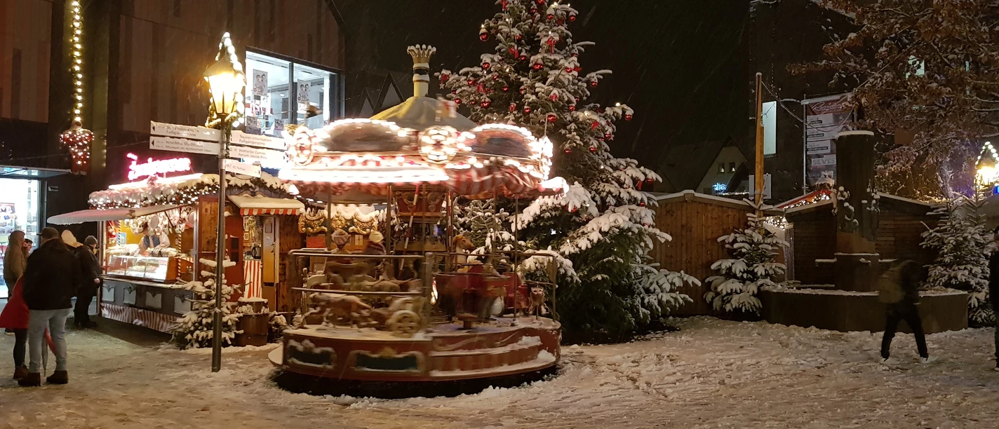 20171210_180126.jpg Ein verschneiter Weihnachtsmarkt am Abend mit beleuchtetem Karussell und geschmücktem Weihnachtsbaum.