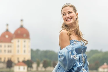 2-Presse_Aschenbrödel_(c)René-Jungnickel.jpg Prinzessin mit blauem Ballkleid lächelt vor märchenhaftem Schloss im Hintergrund.