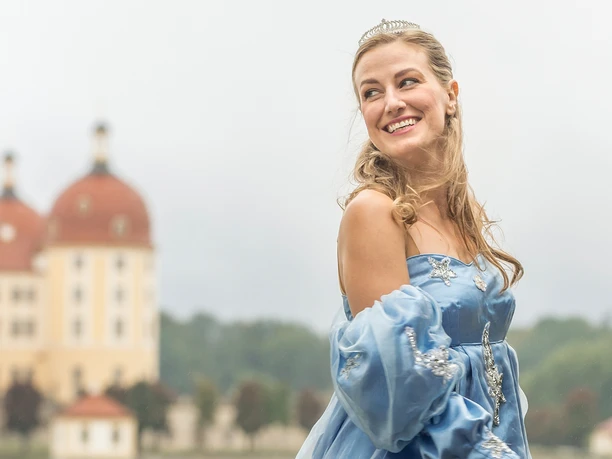 2-Presse_Aschenbrödel_(c)René-Jungnickel.jpg Prinzessin mit blauem Ballkleid lächelt vor märchenhaftem Schloss im Hintergrund.