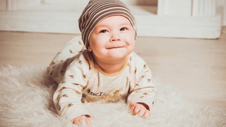 Rosen Photography und Herzenskind.png Ein lächelndes Baby in gestreifter Mütze liegt auf einem flauschigen, weißen Teppich.