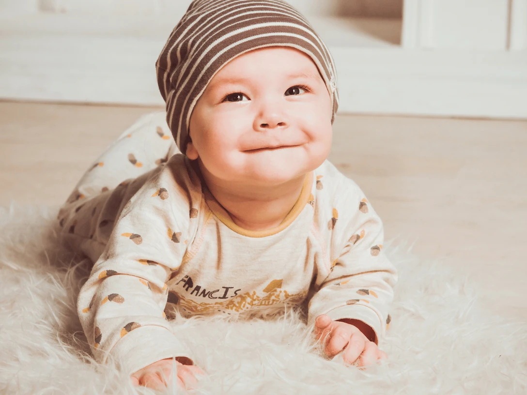 Rosen Photography und Herzenskind.png Ein lächelndes Baby in gestreifter Mütze liegt auf einem flauschigen, weißen Teppich.