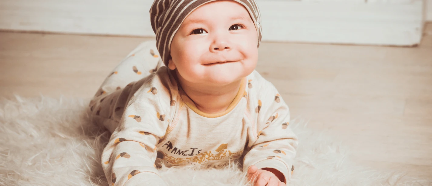 Rosen Photography und Herzenskind.png Ein lächelndes Baby in gestreifter Mütze liegt auf einem flauschigen, weißen Teppich.