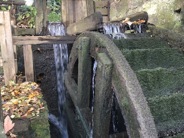 Historische Wassermühle mit hölzernem Wasserrad, umgeben von herbstlichem Laub und fließendem Wasser.