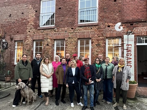 unikat_2024-900010391-22700-11.webp Gruppe von 15 Personen steht vor einem Backsteingebäude, eine Frau hat einen Hund an der Leine.