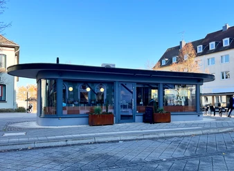 Außenansicht, "Caffé Santa Casa" in Heiligenhaus Ein modernes, rundes Café mit großen Fenstern und hölzernen Pflanzkästen vor der Fassade.